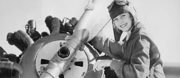 black and white image of a woman in historical pilot clothes, tuning the engine of an airplane